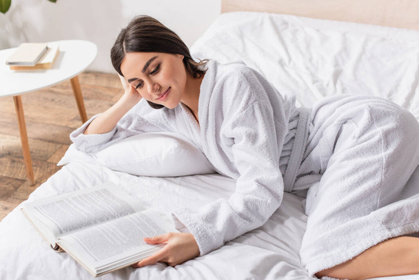 young brunette woman in bathrobe lying on bed and smiling while reading book - Foto, afbeelding