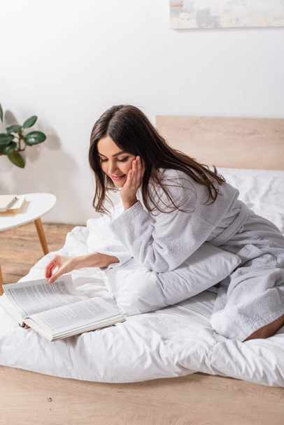 brunette woman in bathrobe sitting on bed and smiling while reading book - Fotó, kép