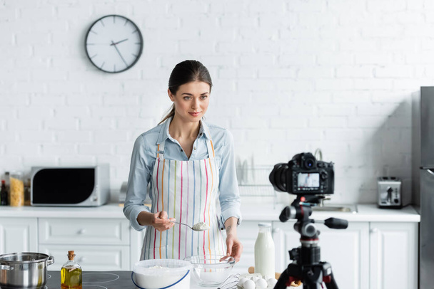 jeune blogueur culinaire tenant cuillère avec de la farine devant un appareil photo numérique flou - Photo, image