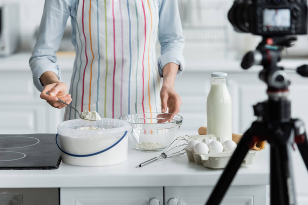 cropped view of woman in apron adding flour into bowl near blurred digital camera - Photo, Image