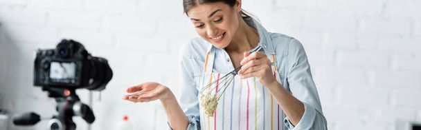 blogueur culinaire joyeux tenant fouet avec pâte crue près de l'appareil photo numérique flou, bannière - Photo, image