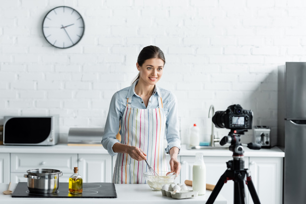 mujer sonriente preparando la masa delante de la cámara digital en la cocina - Foto, Imagen