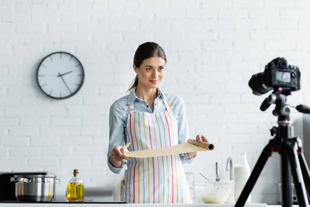 kulinářský blogger držící pekařský pergamen u mísy s těstem a rozmazaným digitálním fotoaparátem - Fotografie, Obrázek