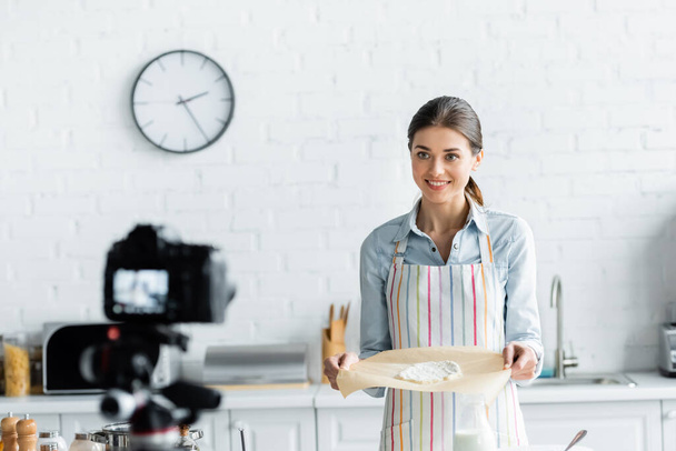 happy culinary blogger showing baking parchment with raw dough near blurred digital camera - Zdjęcie, obraz