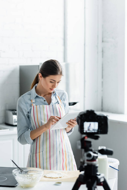 young housewife using digital tablet near raw dough and blurred digital camera - Foto, afbeelding
