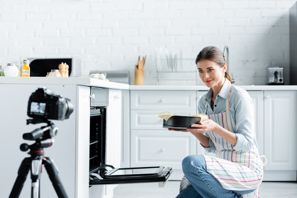 blurred digital camera near young culinary blogger holding baking form near oven - Photo, Image