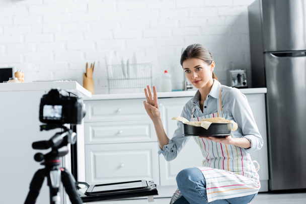 kulinářský blogger ukazující čas vaření při držení formy pečení v blízkosti rozmazaného digitálního fotoaparátu - Fotografie, Obrázek