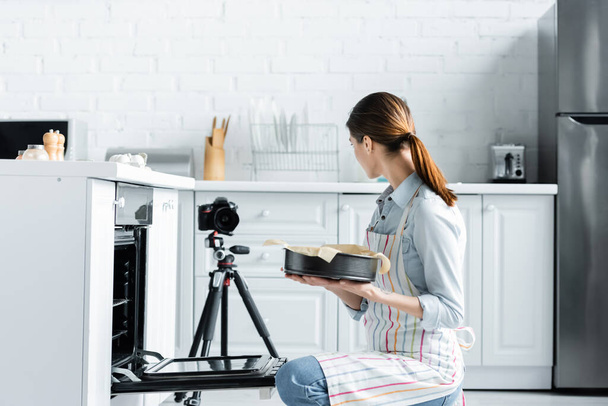 młody gospodyni gospodarstwa domowego gospodarstwa formy do pieczenia w pobliżu piekarnika i aparat cyfrowy w kuchni - Zdjęcie, obraz