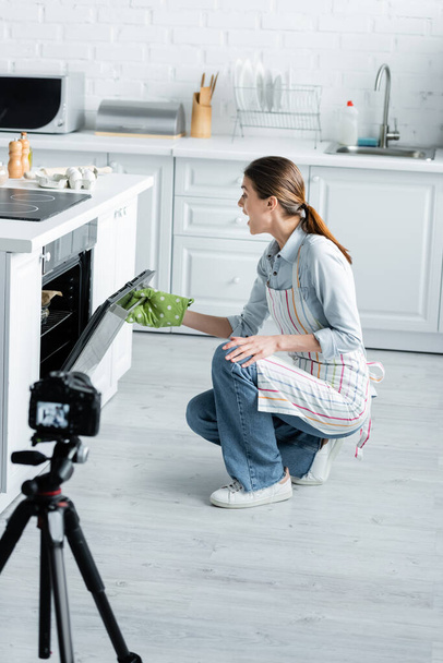 amazed culinary blogger opening oven near blurred digital camera in kitchen - Valokuva, kuva
