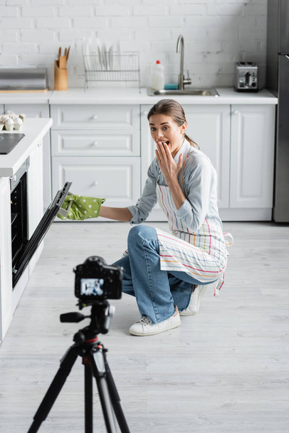 verblüffter kulinarischer Blogger deckt Mund zu, während er Ofen in der Nähe verschwommener Digitalkamera öffnet - Foto, Bild
