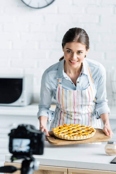 blurred digital camera near happy culinary blogger holding tasty pie - Фото, изображение