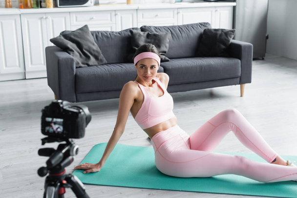 young woman in sportswear sitting on fitness mat near blurred digital camera - Foto, imagen