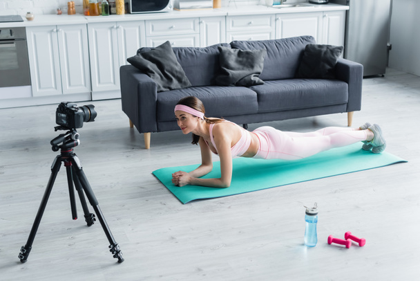 pretty sports blogger looking at digital camera while exercising in plank pose on fitness mat - Foto, Imagem