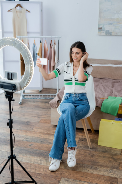 blogger de moda bonita y positiva tomando selfie cerca de la ropa y el titular del teléfono con lámpara de círculo - Foto, Imagen