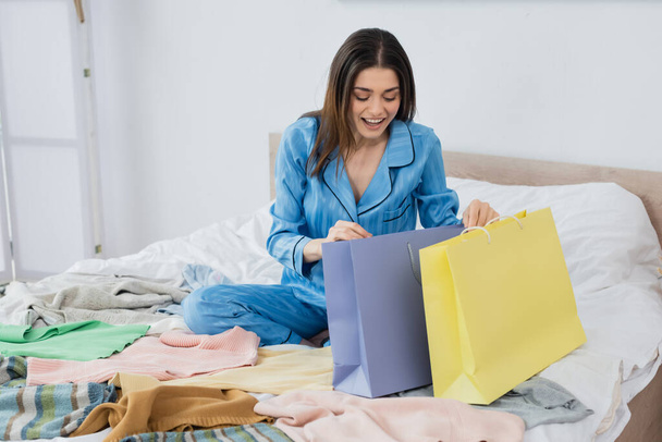 cheerful woman looking into shopping bags near plenty of clothing in bedroom - Foto, afbeelding