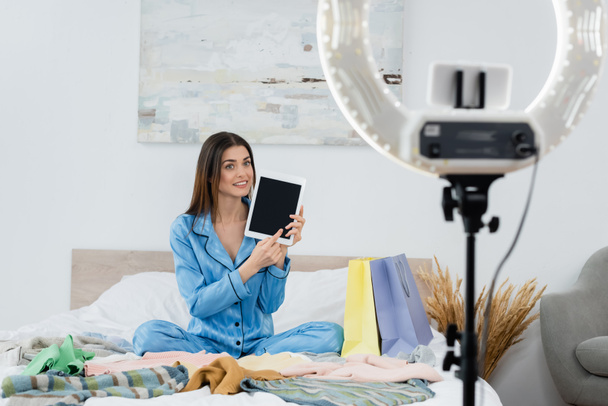 gelukkig vrouw in zijde pyjama wijzend op digitale tablet met leeg scherm in de buurt van kleding en wazig telefoon houder - Foto, afbeelding