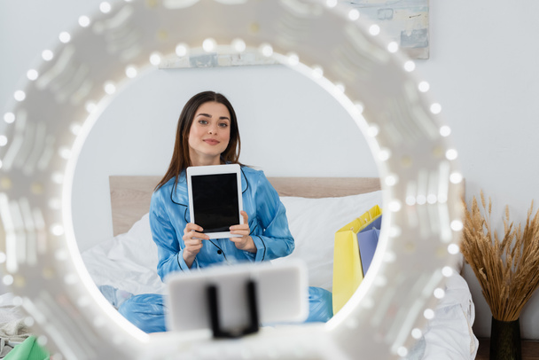 blurred phone holder with ring lamp near fashion blogger in stylish pajama holding digital tablet  - Fotó, kép