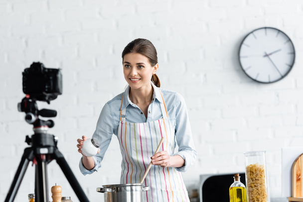 mujer sonriente agregando sal en una cacerola frente a una cámara digital borrosa en la cocina - Foto, imagen