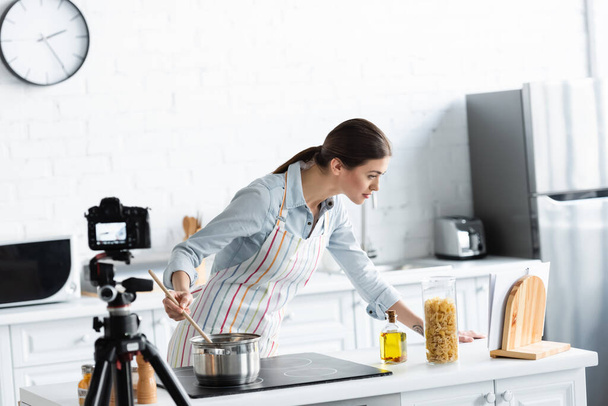 młoda kobieta patrząc na książkę kucharską podczas przygotowywania żywności przed zamazanym aparatem cyfrowym - Zdjęcie, obraz