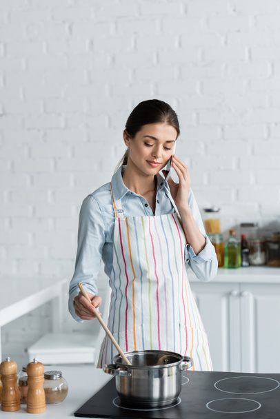 pretty housewife talking on mobile phone while preparing food in kitchen - Foto, Bild