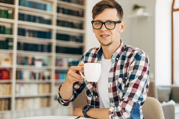 young student in glasses and a plaid shirt drinks coffee and relaxes sitting in the university library against the background of the wall with books. - Foto, Bild