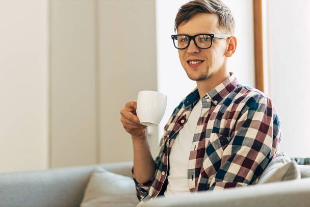 Happy young business man in glasses drinking aromatic coffee from a cup sitting in a cafe near the window - 写真・画像