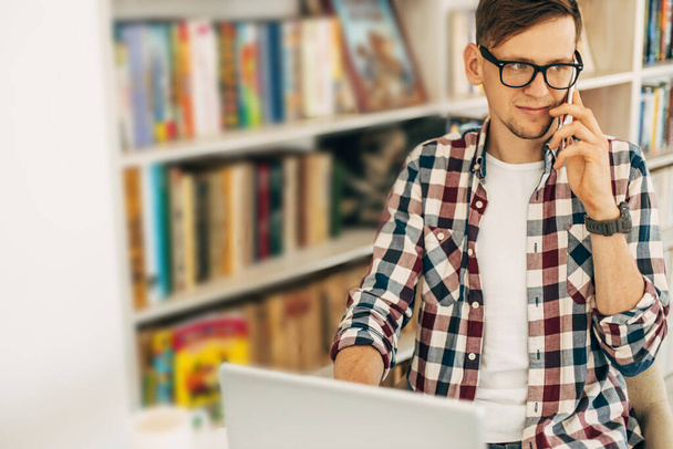 Smiling young stylish male student wearing glasses, successful man using laptop and talking on mobile phone, student studying against the background of the wall with books in the library - Foto, Bild
