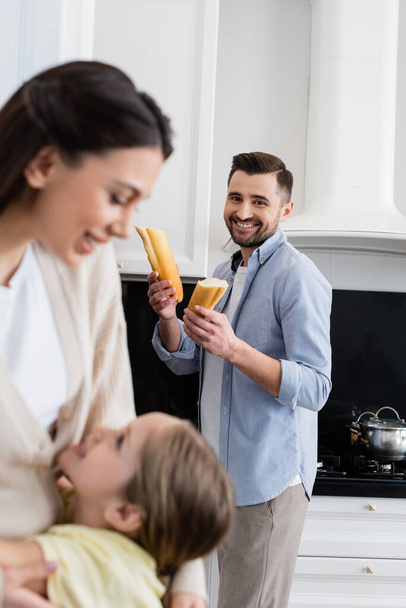 feliz hombre sosteniendo el pan cerca borrosa esposa e hija abrazando en cocina - Foto, Imagen