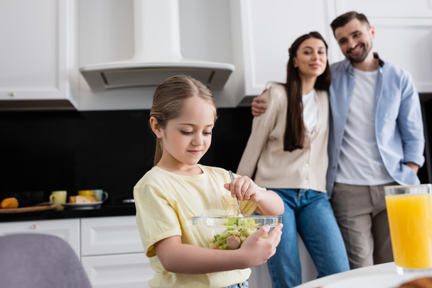girl mixing vegetable salad near happy parents on blurred background - Photo, Image
