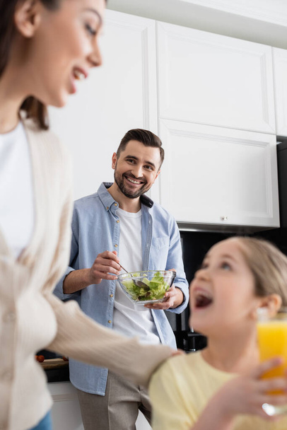 smiling man mixing vegetable salad near blurred wife and daughter in kitchen - Foto, Imagem