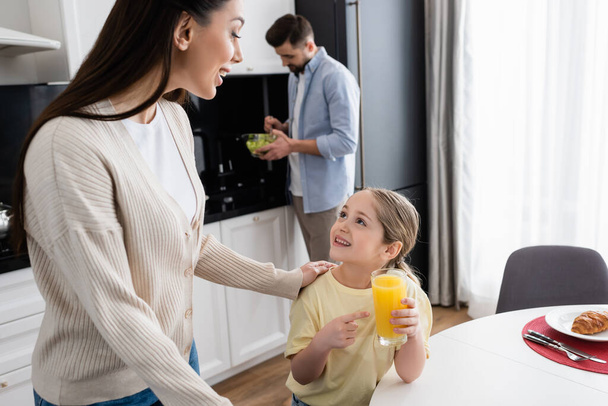 cheerful girl pointing at orange juice near mom and dad mixing vegetable salad on blurred  background - Фото, зображення