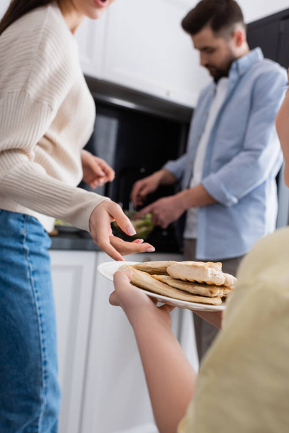 meisje holding plaat met gebakken kip filet in de buurt wazig ouders bereiden ontbijt - Foto, afbeelding