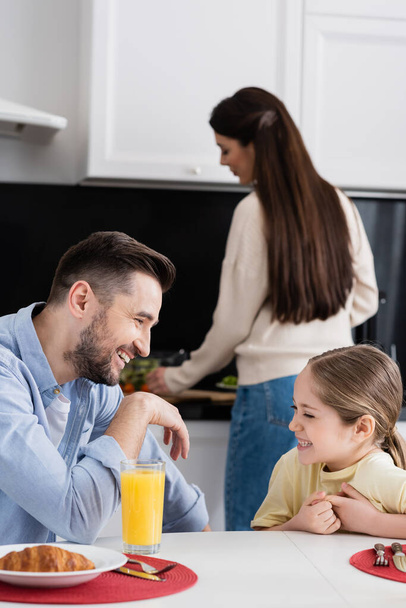 happy father and daughter laughing during breakfast in kitchen near blurred mother - Photo, Image