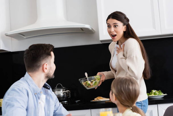 amazed woman touching chest while holding vegetable salad near blurred family - Φωτογραφία, εικόνα