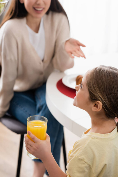 cheerful girl holding glass of orange juice near blurred mother pointing with hand - Photo, Image