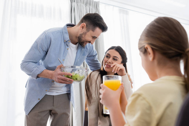 happy man embracing wife while holding bowl with salad near blurred daughter with orange juice - Photo, Image