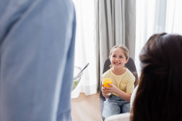 cheerful girl holding glass of orange juice near blurred parents - Foto, afbeelding