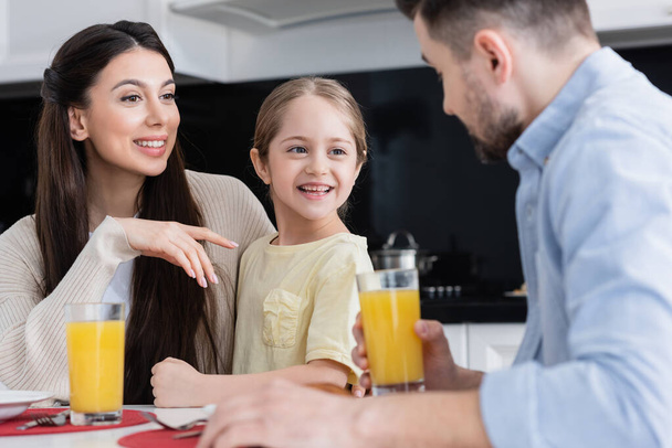 happy woman pointing at cheerful daughter near blurred husband with glass of orange juice - Fotó, kép