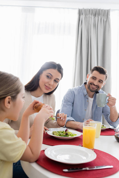 girl eating croissant near happy parents during breakfast - Фото, зображення