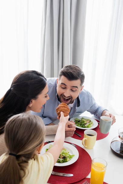 girl feeding father with croissant near happy mom during breakfast - Photo, Image