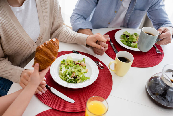 cropped view of girl holding croissant near parents and fresh vegetable salad - Фото, зображення
