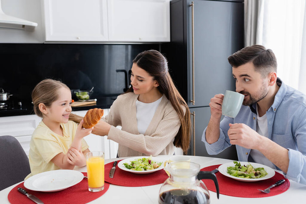 cheerful girl holding croissant near parents and vegetable salad during breakfast - Photo, Image