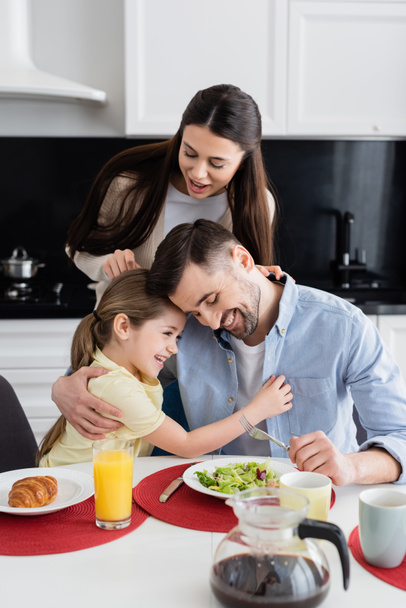smiling woman standing near happy husband and daughter embracing during breakfast  - Photo, Image