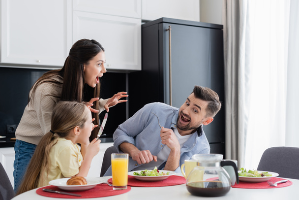 woman scaring excited husband and daughter having breakfast in kitchen - Fotoğraf, Görsel