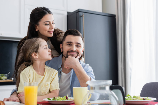 happy man looking at camera near wife and daughter during breakfast - Foto, Imagen