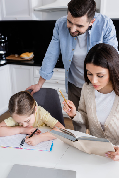 smiling man standing near wife reading book near daughter writing in notebook in kitchen - Foto, imagen