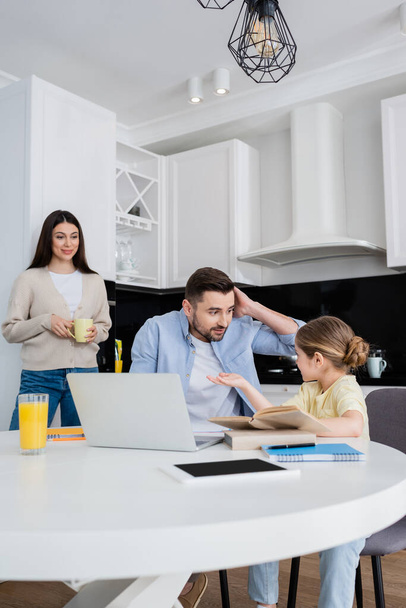 girl pointing with hand while doing homework with confused father near smiling mom - Foto, Bild