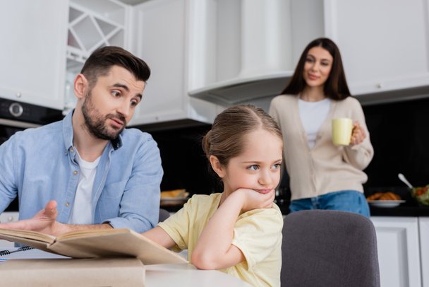 muž mluví s uraženou dceru, zatímco dělá domácí úkoly v blízkosti manželky na rozmazaném pozadí - Fotografie, Obrázek