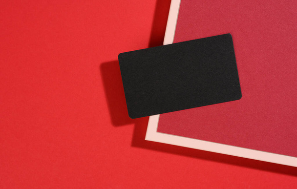 blanco zwart rechthoekig visitekaartje ligt op een moderne rode achtergrond met rode vellen papier met een schaduw. Bedrijfssjabloon, plat lay - Foto, afbeelding