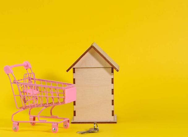 黄色の背景に木造住宅と金属製のミニチュアカート。不動産購入の概念、住宅ローン - 写真・画像